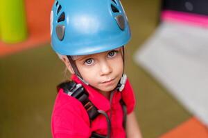 fofa pequeno menina alpinista dentro azul protetora capacete e engrenagem para escalada em pé dentro alpinista Centro diversão parque para crianças foto