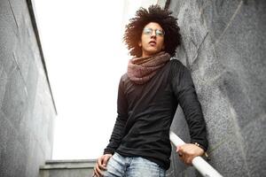 bonito jovem homem do misturado raça com uma corte de cabelo dentro elegante roupas, jeans e uma cachecol posando contra a fundo do uma parede e escadas fez do cerâmico granito azulejos foto