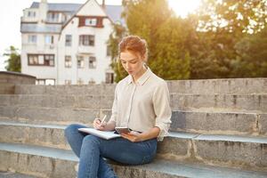 jovem lindo aluna escreve a redação dentro dela caderno sentado em a passos escadas ao ar livre ruivo menina com sardas foto