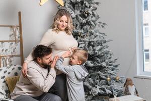 Natal família felicidade retrato do pai, grávida mãe e pequeno filho sentado poltrona às casa perto Natal árvore abraço sorrir foto