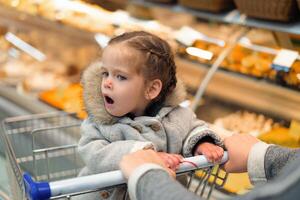 pequeno menina aberto dela boca dentro surpresa enquanto sentado dentro uma supermercado carrinho foto