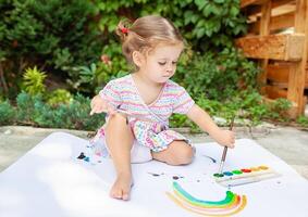 fofa pequeno caucasiano menina desfrutando pintura às a quintal com papel, água cor e arte escovar. seletivo foco foto