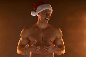 retrato do muscular homem vestindo Natal santa chapéu, mostrando polegar acima em esfumaçado fundo foto