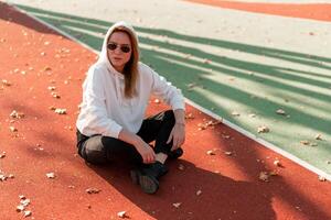 ao ar livre retrato do jovem lindo mulher com grandes dentro oculos de sol e uma branco encapuzado suéter sentado em a quadra esportiva rastrear foto