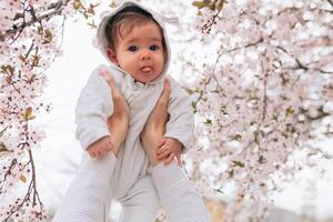 retrato do feliz alegre criança dentro branco roupas sobre árvore flores Flor fundo. família jogando juntos fora. mãe alegremente aguarde pequeno filha foto
