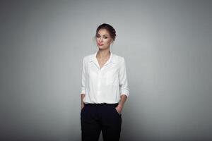 interessado empresária com na moda Maquiagem posando em cinzento fundo dentro estúdio. interior foto do sério jovem senhora dentro branco blusas clássico Preto calça em pé dentro confiante pose.