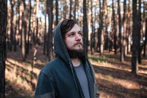 fechar acima retrato do uma barbudo hipster turista dentro cinzento de capuz homem dentro a luz solar madeiras floresta foto