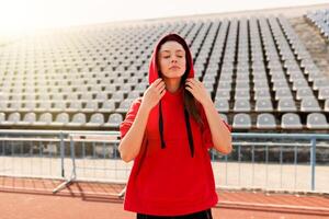 lindo senhora corredor dentro caloroso roupas em a estádio olhando aparte. vestindo vermelho capuz. foto