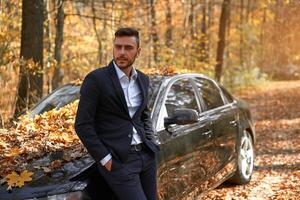 uma bonito jovem caucasiano homem vestindo o negócio terno em pé perto Preto carro em natureza. foto