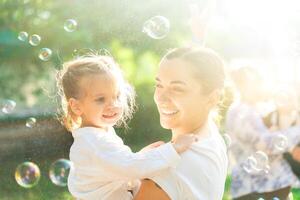 pequeno adorável menina e dela mãe tendo Diversão com Sabonete bolhas em borrado natureza fundo. ao ar livre às a dia com brilhante luz solar. foto