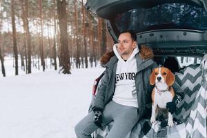 uma jovem família do Viajantes uma cara estão sentado dentro a tronco do seus carro dentro a abraço com seus amigo cachorro beagle. inverno floresta em a fundo. viajando com uma cachorro de carro foto