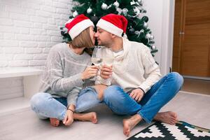 jovem feliz casal dentro Natal chapéus perto uma Natal árvore se beijando, segurando óculos do vinho. Novo ano celebração foto