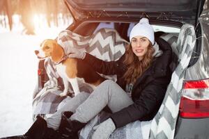 uma jovem família do Viajantes mulher estão sentado dentro a tronco do seus carro dentro a abraço com seus amigo cachorro beagle. inverno floresta em a fundo. viajando com uma cachorro de carro foto
