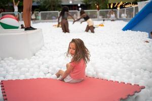 feliz pequeno menina jogando branco plástico bolas piscina dentro diversão parque. foto