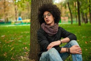 triste afro americano cara com óculos senta dentro a parque em a Relva e parece para dentro a Câmera. à moda aluna com afro corte de cabelo foto