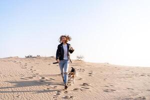 uma jovem caucasiano menina em algum lugar dentro uma couro Jaqueta e azul jeans corre ao longo a arenoso de praia com dela beagle cachorro foto