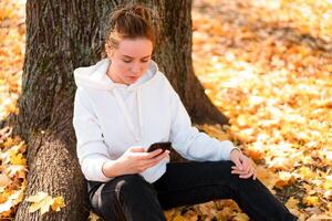 mulher dentro uma branco suéter com uma de capuz senta em terra dentro a parque perto a árvore e segurando uma celular dentro dela mãos foto
