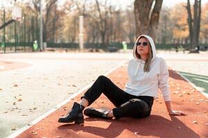 ao ar livre retrato do jovem lindo mulher com grandes dentro oculos de sol e uma branco encapuzado suéter sentado em a quadra esportiva rastrear foto