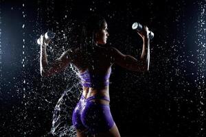 lindo jovem menina dentro roxa roupa de esporte poses com halteres dentro aqua estúdio. gotas do água espalhar sobre dela ginástica corpo. a perfeito figura em a fundo do água salpicos foto