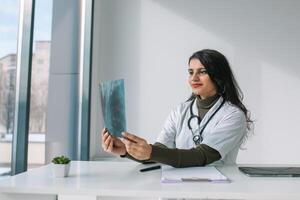 indiano fêmea médico dentro branco casaco com estetoscópio examina raio X e faz notas. jovem indiano médico aluna às a clínica dentro prática foto