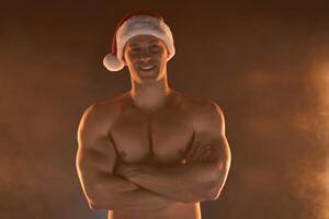 retrato do muscular homem vestindo Natal santa chapéu, guardada mãos em esfumaçado fundo foto