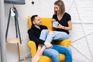 retrato do casal jovem dentro uma acolhedor sala. uma homem senta em uma amarelo cadeira. uma mulher senta em a colo do uma homem e detém uma presente foto