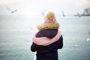 1 menina dentro inverno roupas estão em pé em a doca e alimentando a gaivotas a partir de seus mãos. inverno mar e pássaros. foto