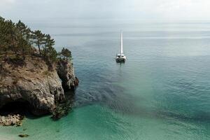 panorâmico Visão do pacífico oceano azul transparente água, uma branco esporte moderno luxo iate vela barco flutuando e uma verde costa com floresta, colinas. feriado de a água e vela corrida. foto