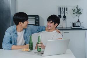 jovem casal gay bebe cerveja enquanto usa o laptop do computador em uma casa moderna. Homens lgbtq asiáticos felizes relaxam a diversão usando tecnologia jogar mídia social juntos enquanto está sentado à mesa na cozinha no conceito de casa. foto