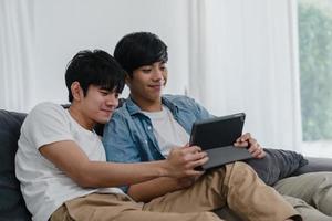 jovem casal gay usando tablet em casa. homens lgbtq asiáticos felizes relaxam a diversão usando a tecnologia, assistindo filme na internet juntos enquanto estava deitado no conceito de sala de estar. foto