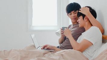 casal de gays asiáticos usando computador laptop e bebendo café em uma casa moderna. jovem amante asiático masculino feliz relaxe descansar juntos depois de acordar, assistindo filme deitado na cama no quarto em casa pela manhã foto