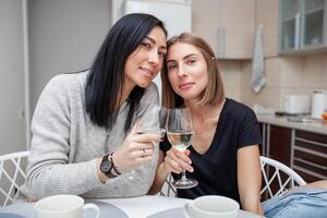 amigos encontro com vinho e bolo dentro a moderno estilo cozinha. jovem mulheres sorrir e Piada com óculos do vinho dentro dele mãos foto