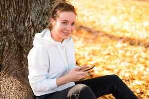 mulher dentro uma branco suéter com uma de capuz senta em terra dentro a parque perto a árvore e segurando uma celular dentro dela mãos foto