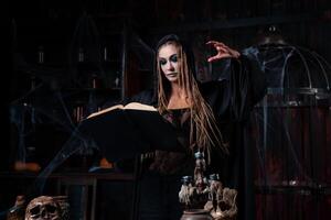 dia das Bruxas conceito. bruxa vestido Preto de capuz com tranças em pé Sombrio masmorra quarto usar Magia livro para conjuração Magia soletrar foto