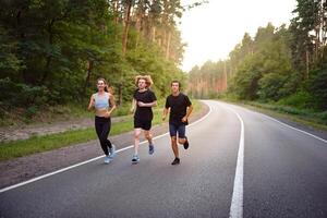uma grupo do três pessoas atletas 1 menina e dois homens corre em a asfalto estrada dentro uma pinho floresta. foto