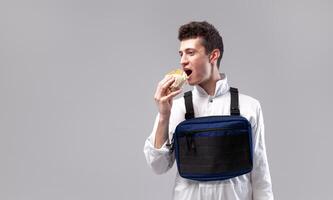 jovem à moda masculino trabalhador com peito equipamento saco é comendo uma saboroso suculento hamburguer em uma branco fundo foto