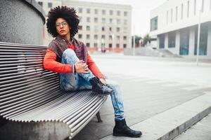 uma bonito jovem egípcio encaracolado cara sentado em uma Banco vestido dentro uma brilhante laranja suéter e jeans. a aluna anda em dentro a pausa entre a aulas. fofa hipster cara foto