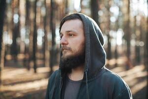 uma jovem homem com uma barba anda em dentro uma pinho floresta. retrato do uma brutal barbudo homem dentro uma de capuz foto