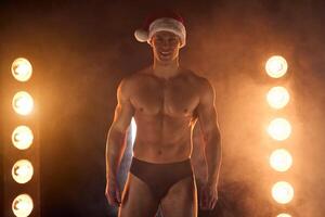 sexy papai noel. muscular cara dentro calça dentro natal chapéu sorridente às Câmera, lâmpadas brilhando em fundo foto