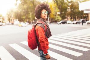 uma jovem africano americano homem com uma vermelho mochila cruzes uma faixa de pedestre dentro uma cidade foto