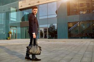 masculino moda. uma jovem masculino homem de negocios dentro uma na moda terno e uma branco camisa com uma grande Preto Bolsa vem Fora a partir de uma elegante roupas loja. foto