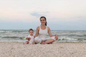 feliz família mãe e criança filha fazendo ioga, meditar dentro lótus posição em de praia às pôr do sol foto