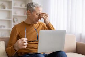 Senior homem fricção olhos enquanto usando computador portátil às casa foto
