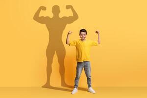 a entusiasmado jovem Garoto dentro uma amarelo camisa flexiona dele braços com confiança foto