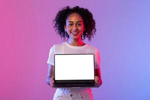 alegre Preto mulher apresentando computador portátil com em branco tela em néon fundo foto