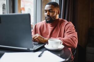 retrato do feliz africano homem de negocios sentado dentro uma cafeteria e trabalhando em computador portátil. foto