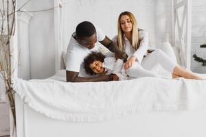 retrato do feliz multirracial jovem família deitado em acolhedor branco cama às lar, sorridente internacional mãe e Papai relaxante com pequeno birracial menina criança posando para cenário dentro quarto foto