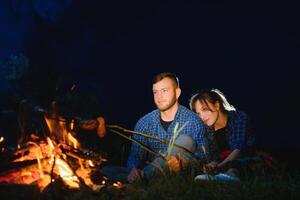 casal cozimento salsichas em a fogo, e relaxante de a fogueira dentro a floresta dentro a noite. romântico fogueira conceito. foto