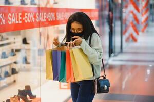 jovem africano americano mulher às compras Shopping dentro Novo normal depois de covid-19 - feliz e lindo Preto menina dentro face mascarar segurando compras bolsas desfrutando às beleza moda loja foto