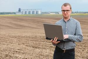 moderno agricultor verificação dele campo plantar e trabalhando em computador portátil computador contra milho secador silos dentro conceito do industrial e agricultura foto